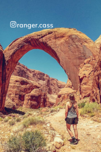 A woman standing in front of a large arch in the desert at Rainbow Bridge National Mountain.